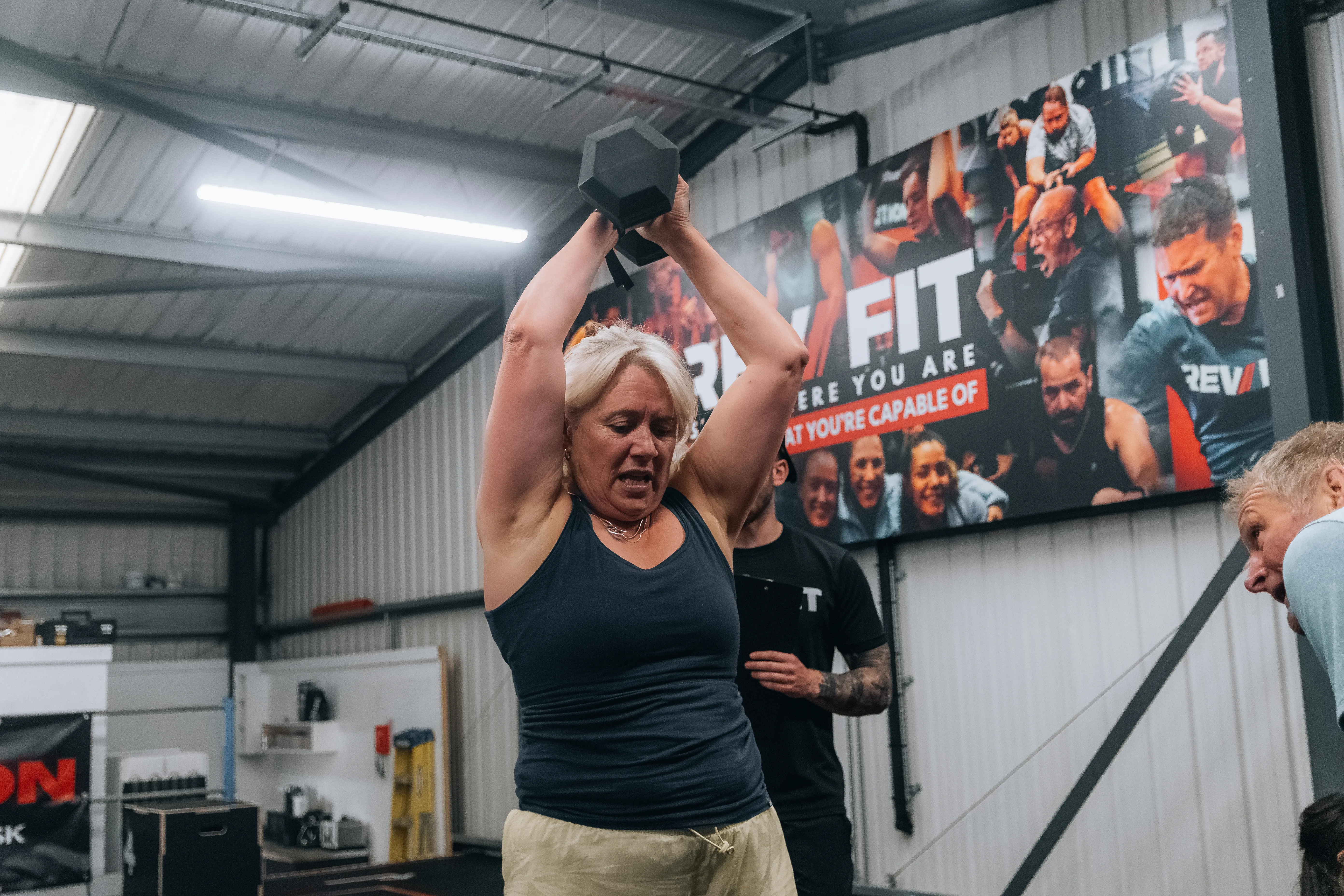 An older competitor pressing a dumbbell overhead in front of the Rev Fit wall.