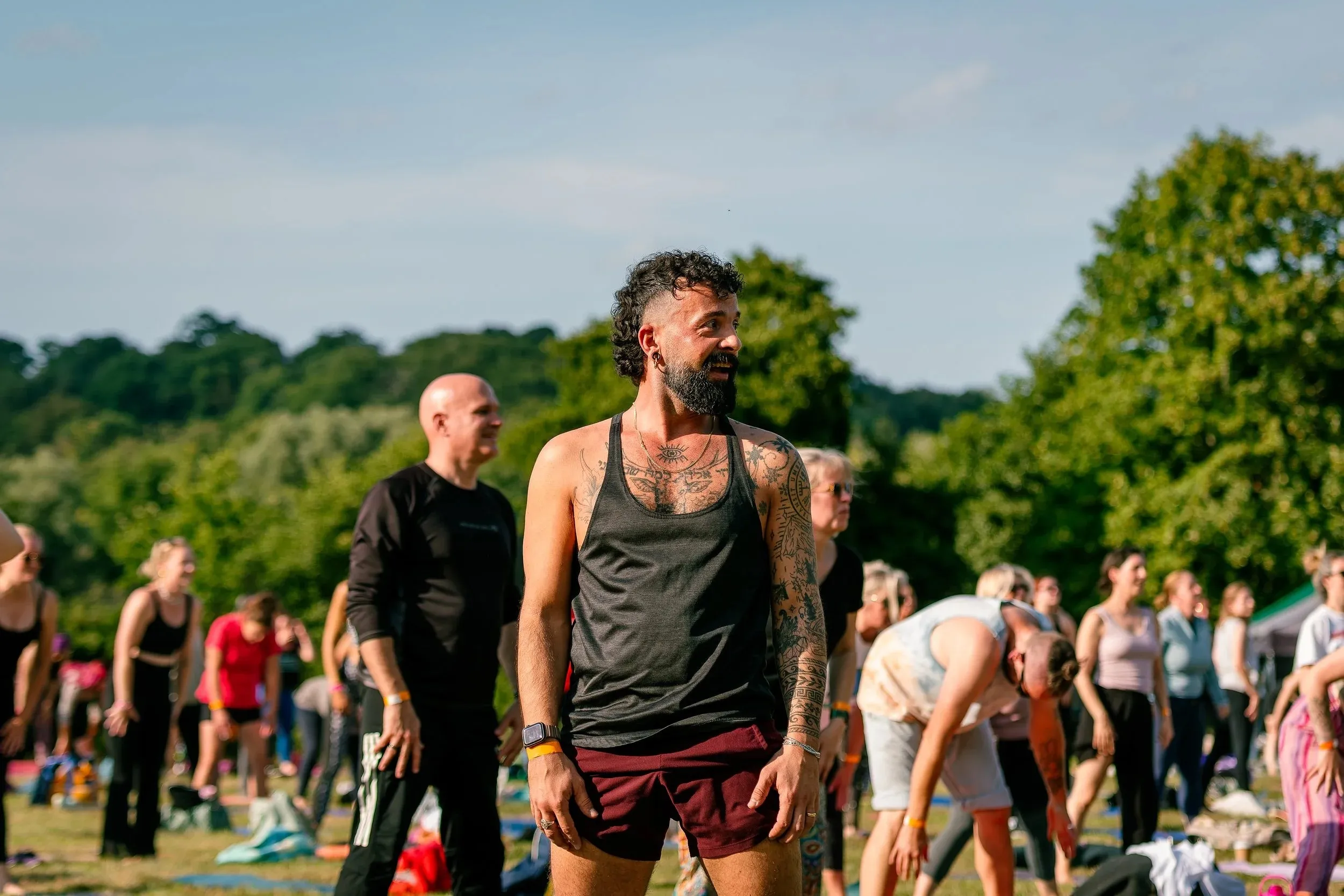 A bearded man in a black vest standing among people warming up on the grass.