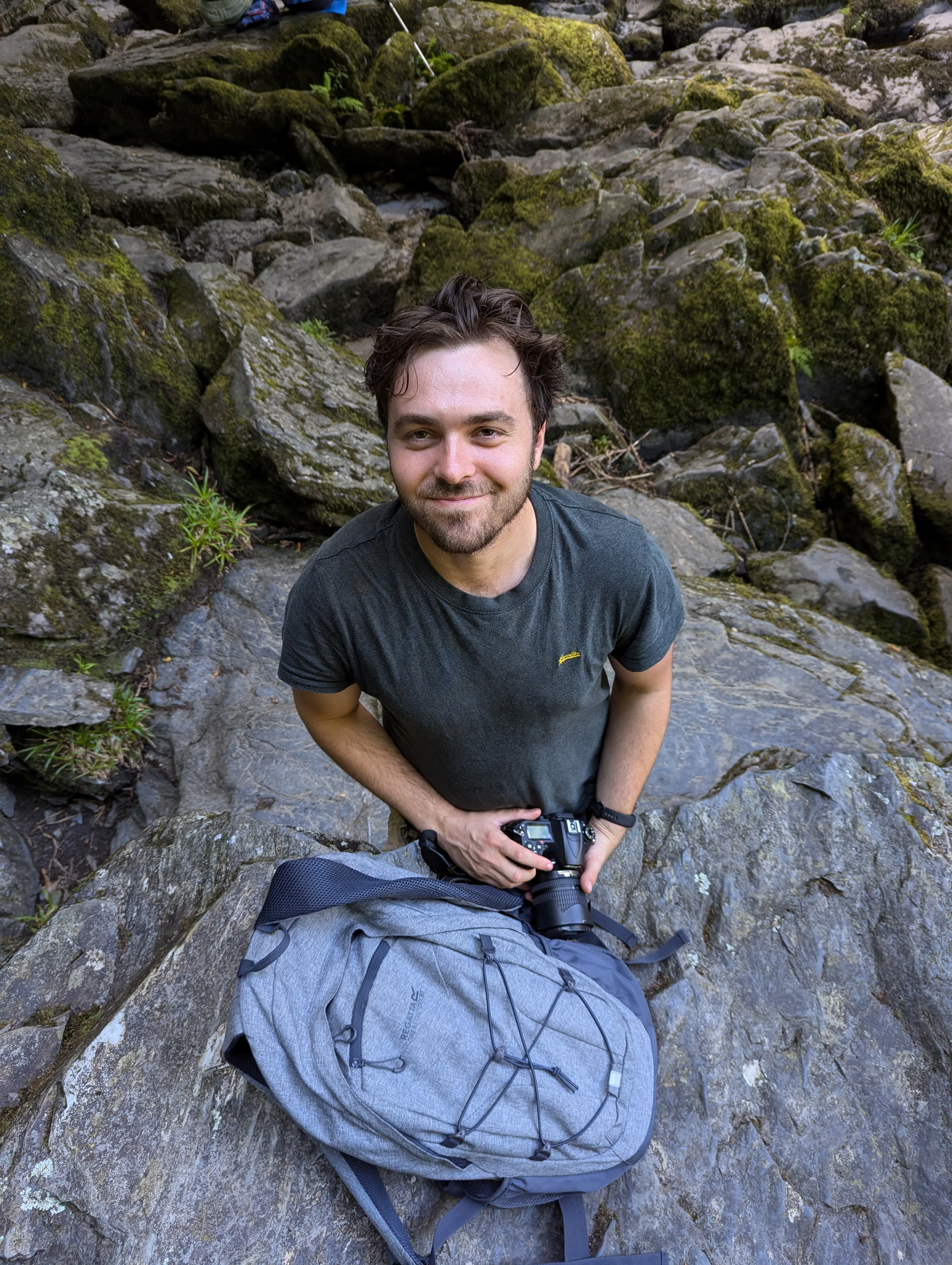 Mike Arnold sitting on mossy rocks with his camera and rucksack, grinning up at the lens.